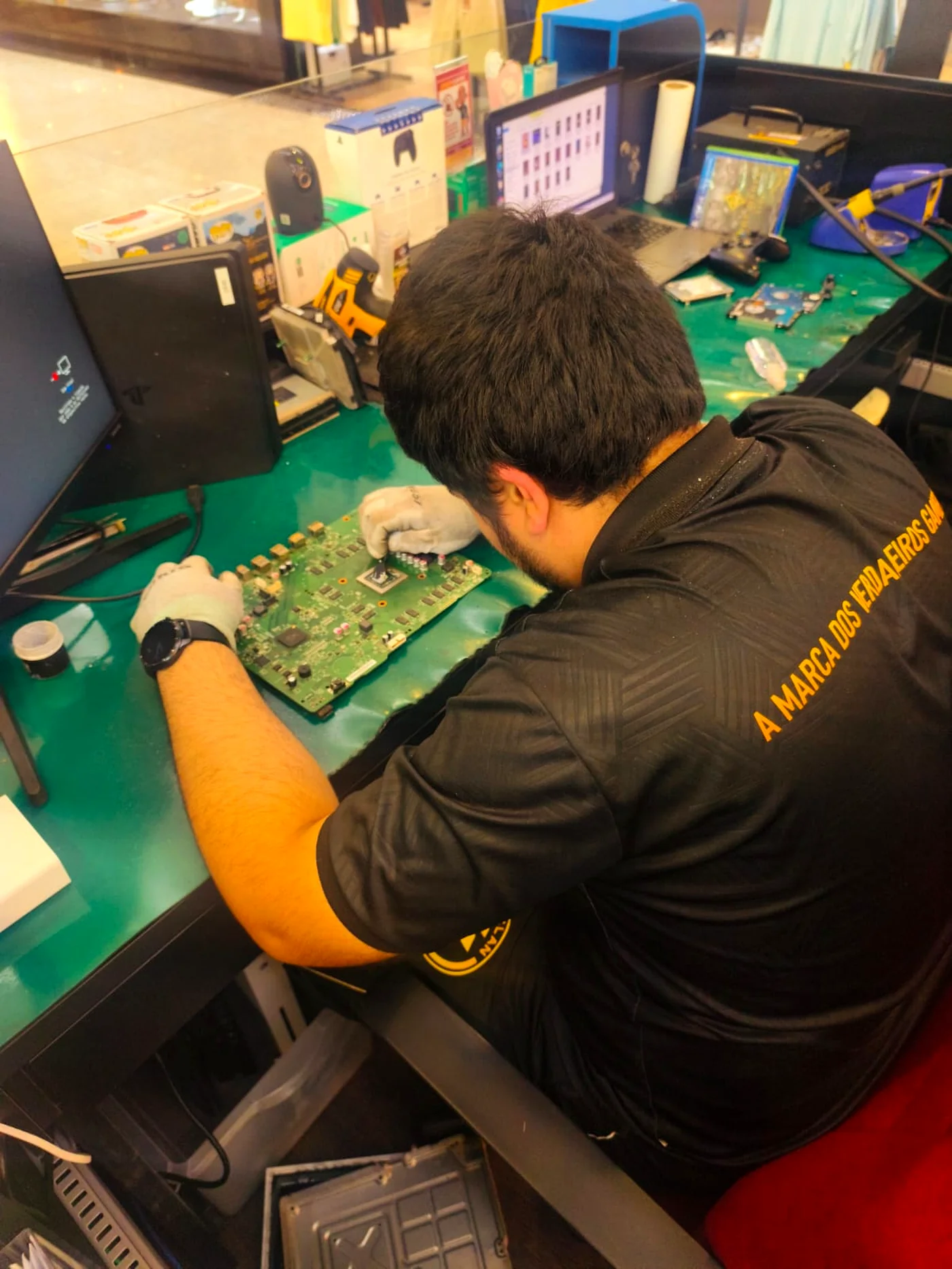 Profissional da CLAN Y GAMER concentrado em manutenção de aparelho eletrônico na bancada do laboratório transparente.
